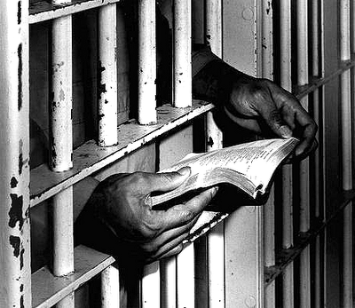 black and white picture with hands holding a book, through prison bars