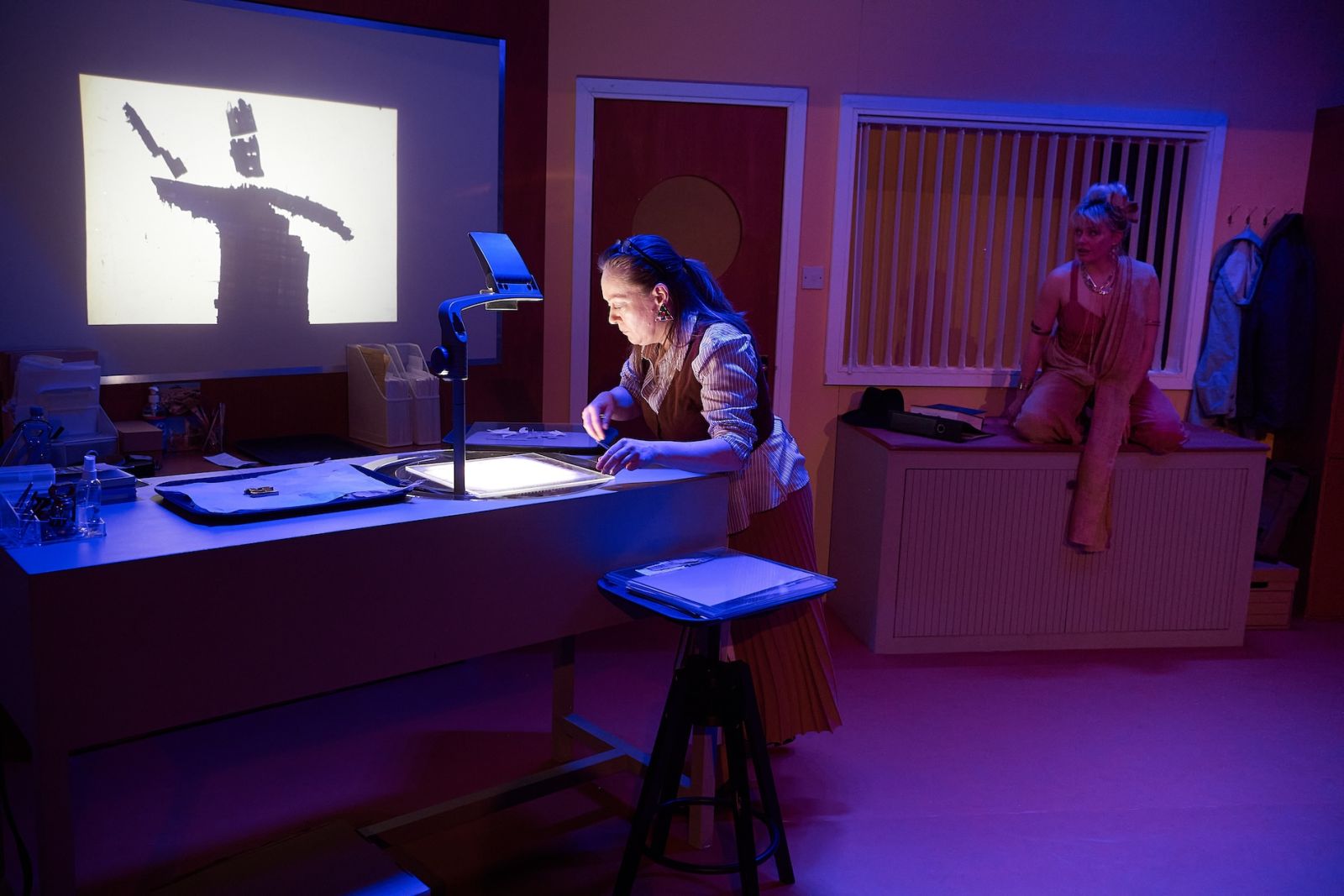 An inside space with overhead projector. A woman peers at the images in the projector, while another woman sits nearby, on a filing cabinet.