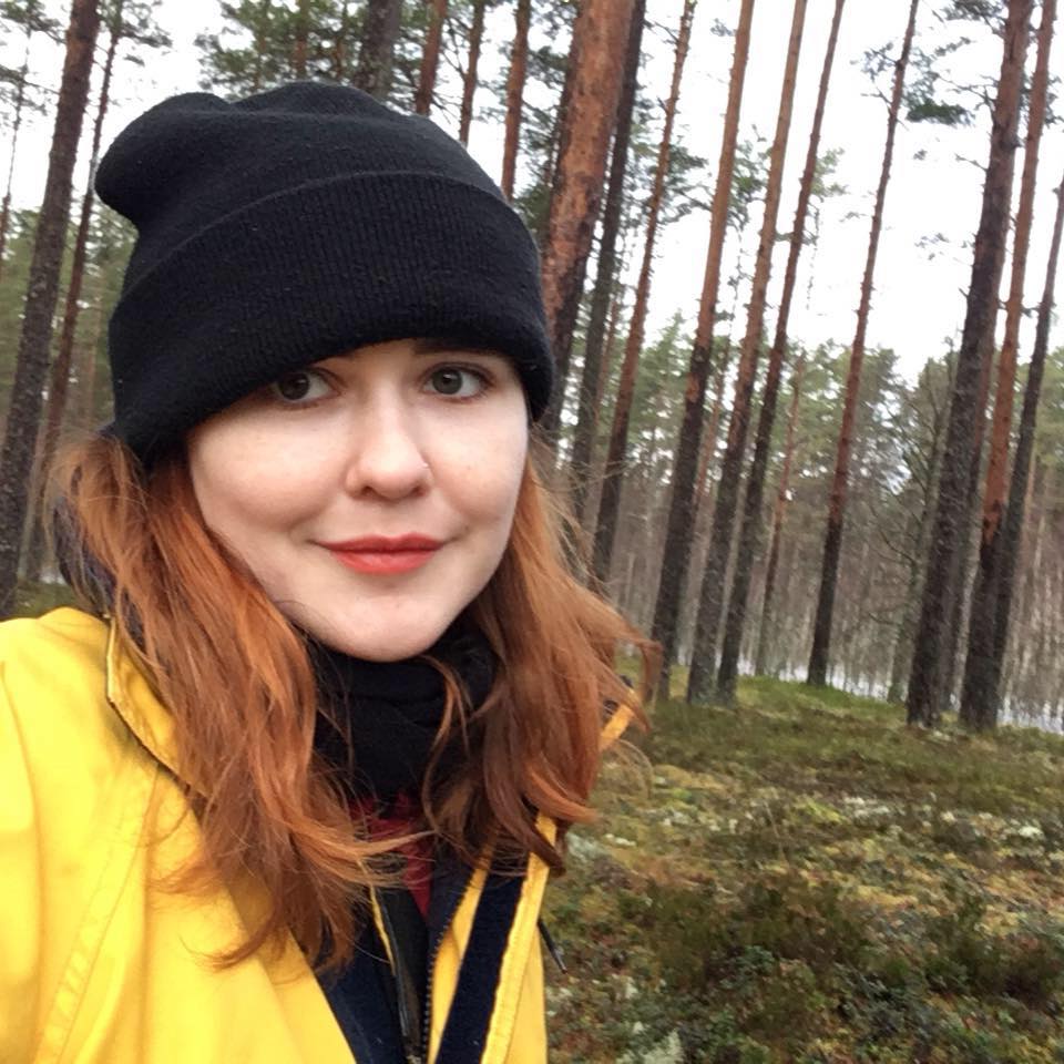 Portrait photo of Katherine Soper wearing a black hat and yellow coat in an outdoor forest landscape