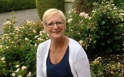 Photo of Peta Wilkinson, Cheif Executive of Willen Hospice, smiling infront of flowers. She has short blonde hair, a white cardigan and funky leopard print glasses on
