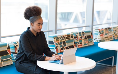 A female with beautiful thick hair, piled up on her head, sits in a black short, typing on her laptop in a room with multi-coloured chairs around her. She looks deep in concentration