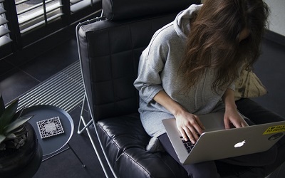 Photo shows a woman with long hair on her laptop, wearing a grey hooded top. She is sitting in a leather chair