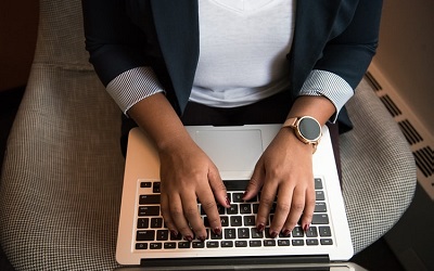Photo of a woman's hands on a laptop, typing. She is wearing a blazer and smart gold watch
