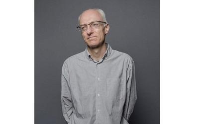 Image shows Dr Rob Macmillan against a grey background. He wears a grey shirt and has glasses. He also has short white hair and is looking away from the camera.