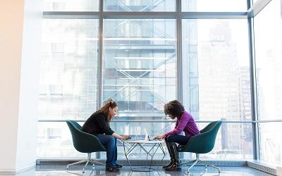 Image shows two females sitting infront of a very large, glass window. They lean over a small table and are deep in concentration. Photo credit WOCinTech available on Unsplash.