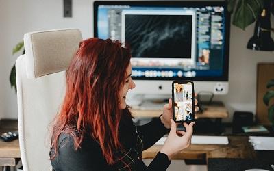 Image shows a young woman with red hair sitting on a large white office chair, holding her phone smiling as she speaks on a live video call. A large computer screen is behind her. She looks very happy and has a nose ring. Photo credit Annie Spratt on Unsplash