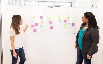 Image shows two women standing at a white board discussing key points that have been shared on post-it notes. Photo credit WOCinTech available on Unsplash