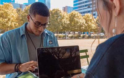 Photo shows a young man in a blue shirt with glasses working on a laptop. He sits opposite a woman who also works on a laptop. There are trees and offices behind them. The image suggests two colleagues are preparing for a meeting. Photo credit Eliott Reyna on Unsplash