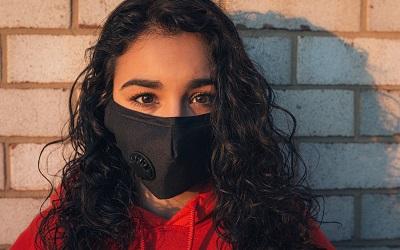 Photo shows a woman with curly, dark hair standing against a brick wall staring into the camera. She has very dark brown eyes and wears a black mask and a red jumper. Photo credit: Gayatri Malhotra, Unsplash