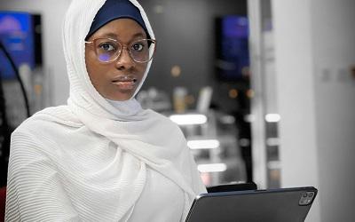 A woman wearing a white hijab sits with a laptop. She is wearing black glasses and staring at the viewer