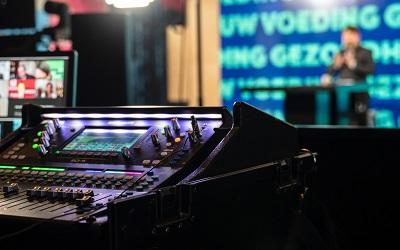 A mixing desk sits next to a screen in front of a stage with a live broadcast taking place