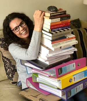 A photo of Michelle sitting on the sofa holding a pile of books and folders as tall as her (topped by a PC mouse).