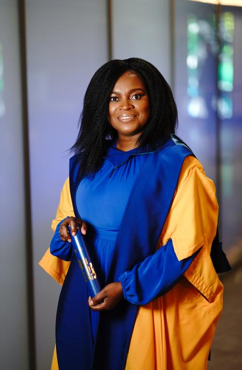 A close-up of Joy-Tendai Kangere holding her honorary degree certificate. She is smiling confidently, wearing the traditional blue and gold graduation gown, representing a moment of achievement and recognition at The Open University's Dublin ceremony.