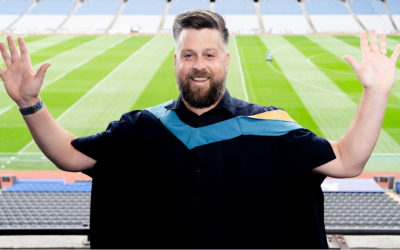 Sean smiling, in academic robes, raises both arms, one hand holding his degree. 