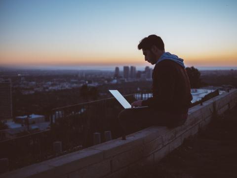 A young man sitting on a wall over looking a city at dusk while using his laptop