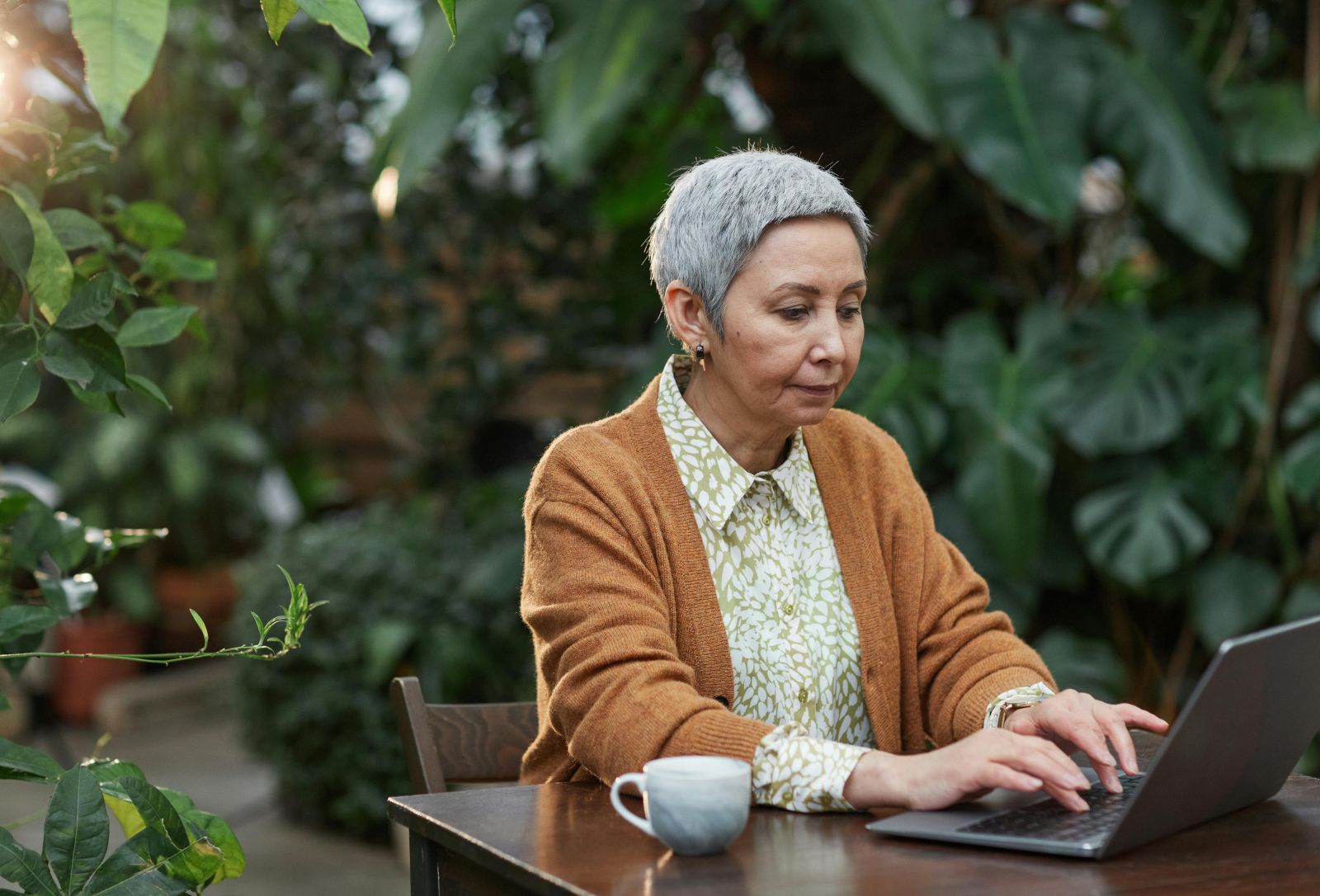 A person sitting at a table using a computer, outside. Photo by Marcus Aurelius: https://www.pexels.com/photo/woman-busy-using-her-laptop-6787902/