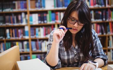 A student is sitting at a table covered in books whilst studying in a library. Rows of bookshelves can be seen behind them.