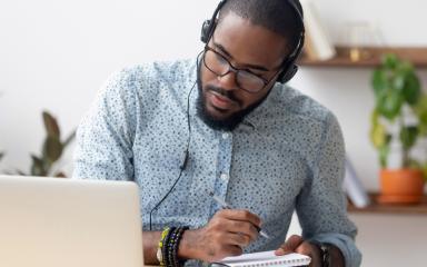 A student is sitting at a desk, studying at home on his laptop. He is wearing headphones and writing notes.