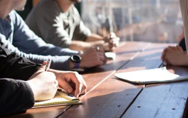 A group of people are sitting at a desk together writing in notepads.