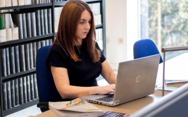 A student is sitting at a desk in a Library which has some textbooks and an open laptop placed upon it. There is a bookshelf behind them.