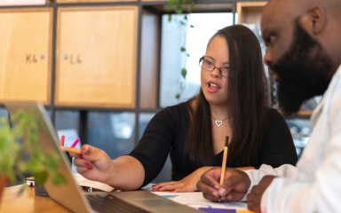 Two business professionals are sitting next to each other looking at an open laptop screen having a discussion.