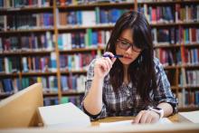 A student is sitting at a desk in a Library with a pen to her mouth, thinking about what to write. Behind her is a large bookshelf.