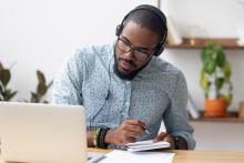 A student is sitting at a desk, studying at home on his laptop. He is wearing headphones and writing notes.