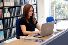 A student is sitting at a desk in a Library which has some textbooks and an open laptop placed upon it. There is a bookshelf behind them.