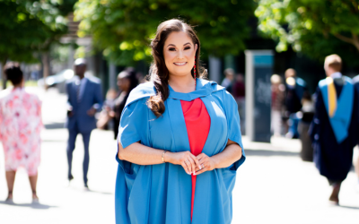 A female student with with brunette hair smiling into the camera while wearing OU degree ceremony robes. She's stood outside in the sun with blurred trees in the background.