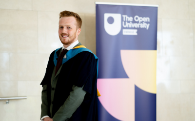 Nick, wearing OU degree ceremony robing, is stood in front of an OU banner stand and smiling into the camera.