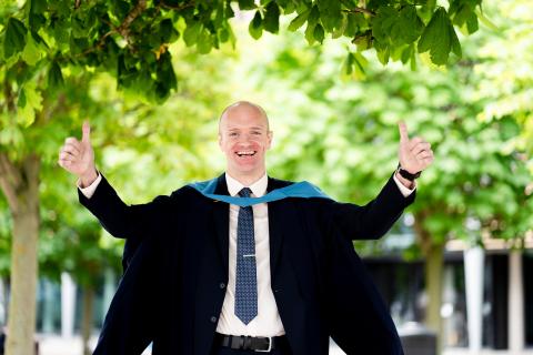 Brendan stood outside by some trees, he is wearing a black suit, Open University degree robes and large smile. Both his arms are raised in celebration.