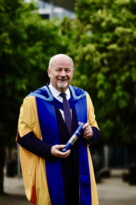 Brian Rowan standing in his Hon Graduate robes with a scroll in his hands