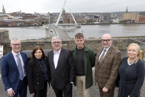 A group of 6 people standing posing for a photo with the peace bridge in Derry/Londonderry in the background