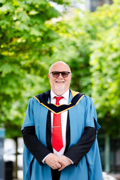 A picture of paul stood in front of some trees outside. He is wearing a red tie and blue OU degree ceremony robes.