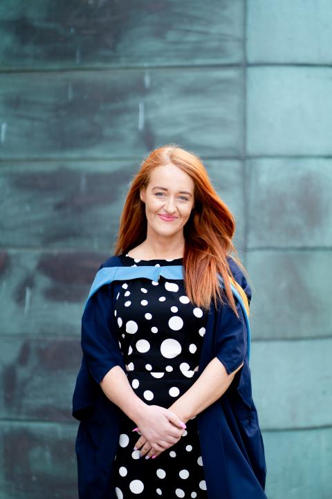 Stacey wearing a spotted dress and OU degree ceremony robes stood in front of a light blue tiled wall