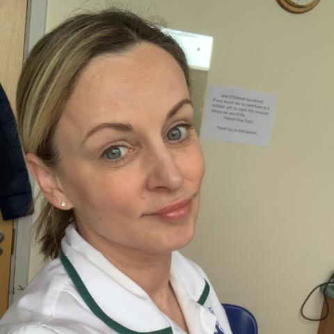 A woman wearing a white uniform with green piping stands indoors, looking at the camera with a gentle smile. Her hair is pulled back, and she has light-colored eyes and subtle makeup.