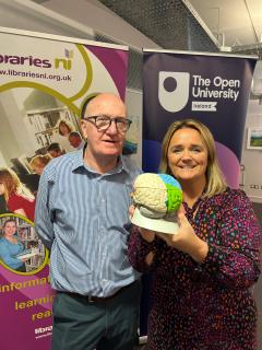 A man and a woman stand in front of banners for Libraries NI and The Open University Ireland. The man, wearing glasses and a blue checkered shirt, stands to the left, while the woman, smiling and holding a colorful model of a brain, is on the right. The woman is wearing a patterned dress with pink, blue, and purple colors. The background features informational material promoting learning and reading at Libraries NI.