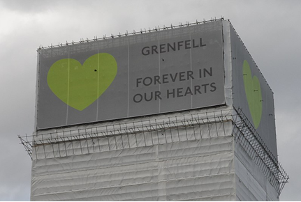 A photo of the structure Grenfell Tower after the fire. A banner is at the top of the tower. The banner has a grey background, with a green heart, and reads 'Grenfell. Forever in our hearts.'