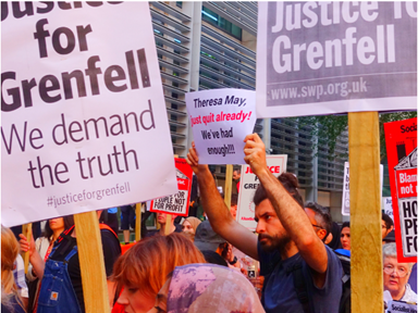 An image of a protest. The protest contains a number of placards and signs. One of the placards says 'Justice for Grenfell, we demand the truth.' A man is holding up a sign which says 'Theresa May just quit already. We've had enough!!!'