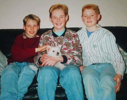 An image of Robbie Powell with his two brothers. They are sat on a sofa together, with a dog on the sofa with them.