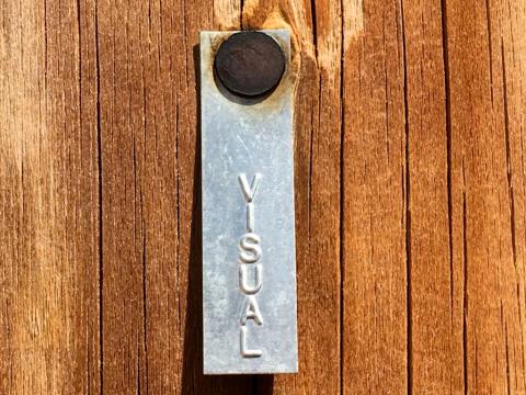 A photograph of the word 'visual' inscried into a small metal tag, nailed into a wooden surface