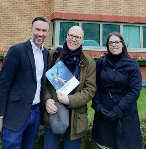 A white man, a white man holding an open university book, and a white woman, all standing in front of a building.