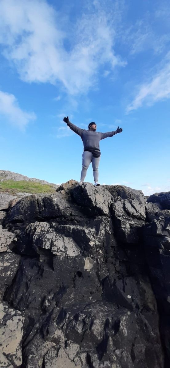 Armando stands on a high rock with his arms stretched wide with the blue expanse of the sky behind him