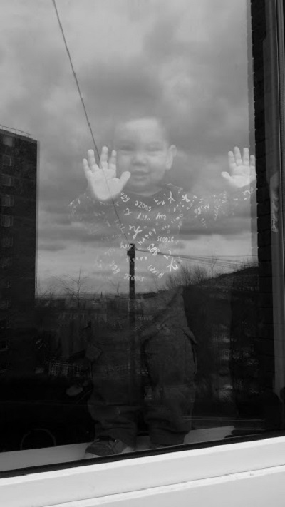 A gorgeous, happy toddler stands with his hands pressed up against a window smiling at the refugee art photographer