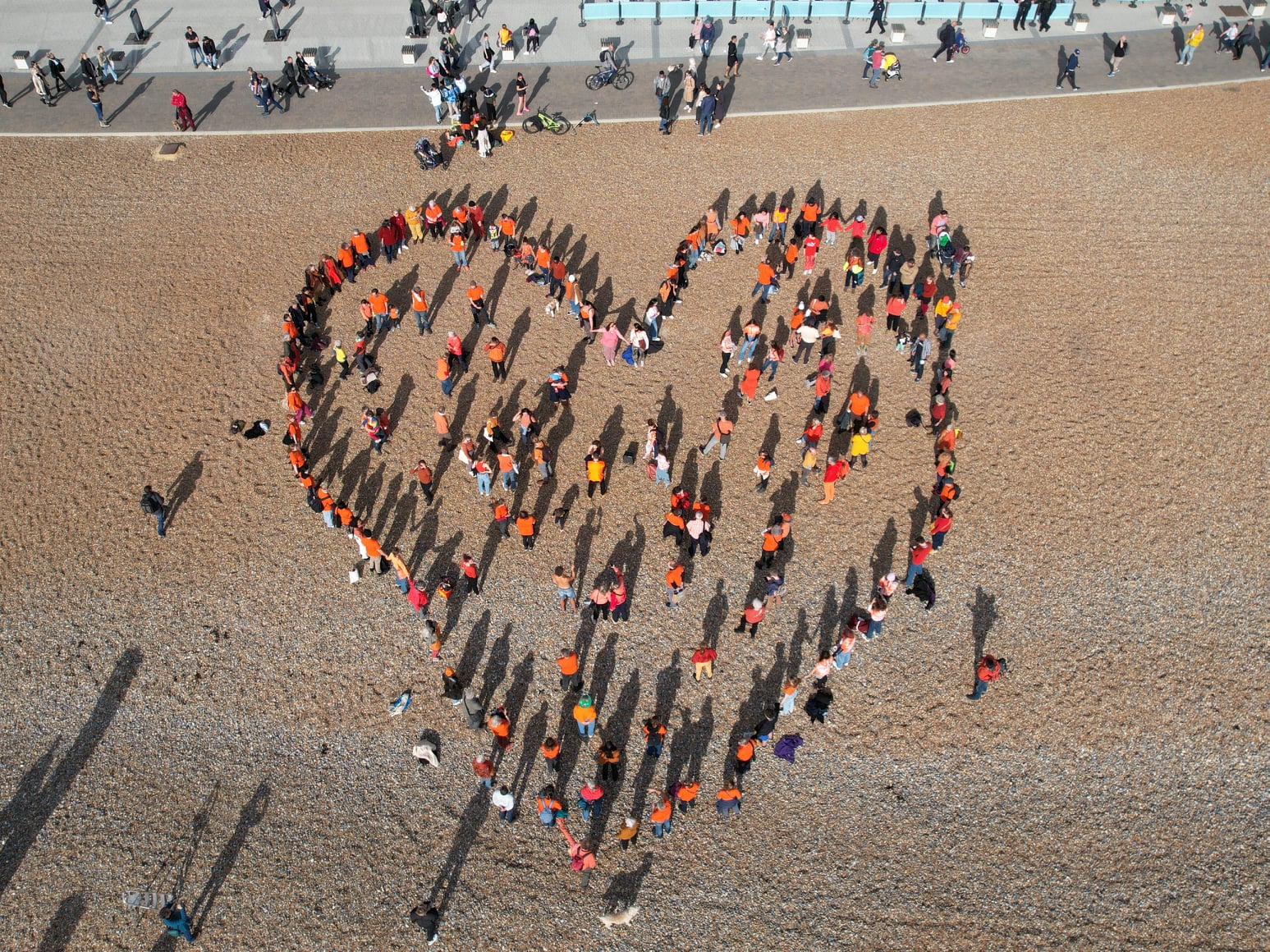 Community in Brighton stood on the beach in the shape of a heart, all wearing orange