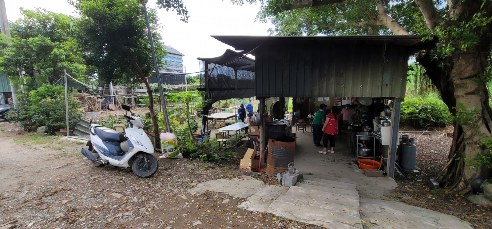 A small piece of farmland in a neighbourhood in Taipei, Taiwan. There is a motorcycle parked in the foreground, and a gazebo to the right. In the distance are some larger concrete buildings.