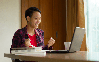 A happy, smiling teenage boy is pictured sitting at a table, with a laptop