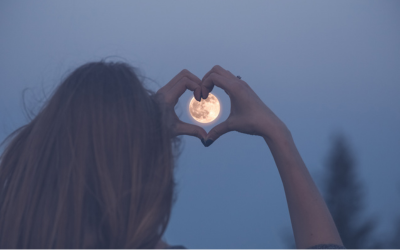 The moon pictured from a distance, with a person using their hands to make a heart shape around it