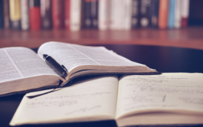 Photo of a notepad, pen and an open book, in front of a bookshelf. By Aaron Burden on Unsplash. 