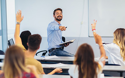 A teacher at the front of a class, pointing to a student who has their hand up